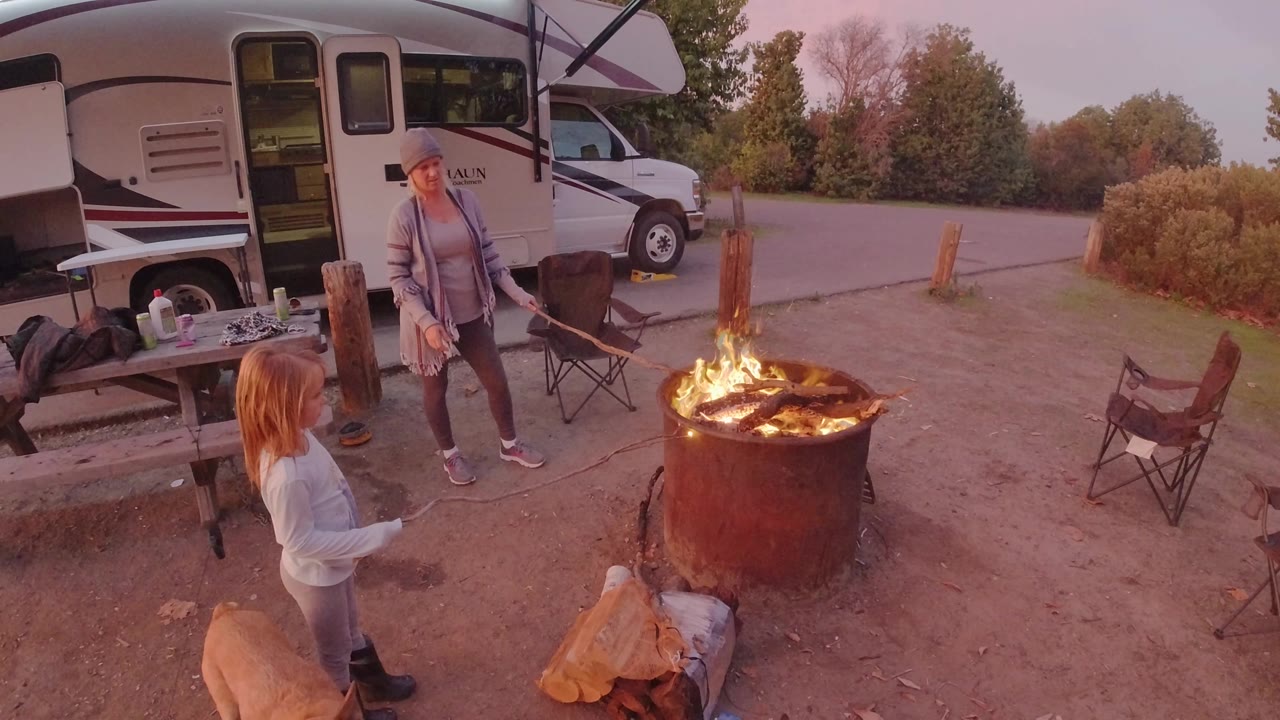 At a San Clemente RV campsite, an adult and a child stand near a large metal fire ring with flames, folding chairs, logs, and a picnic table in warm evening light.