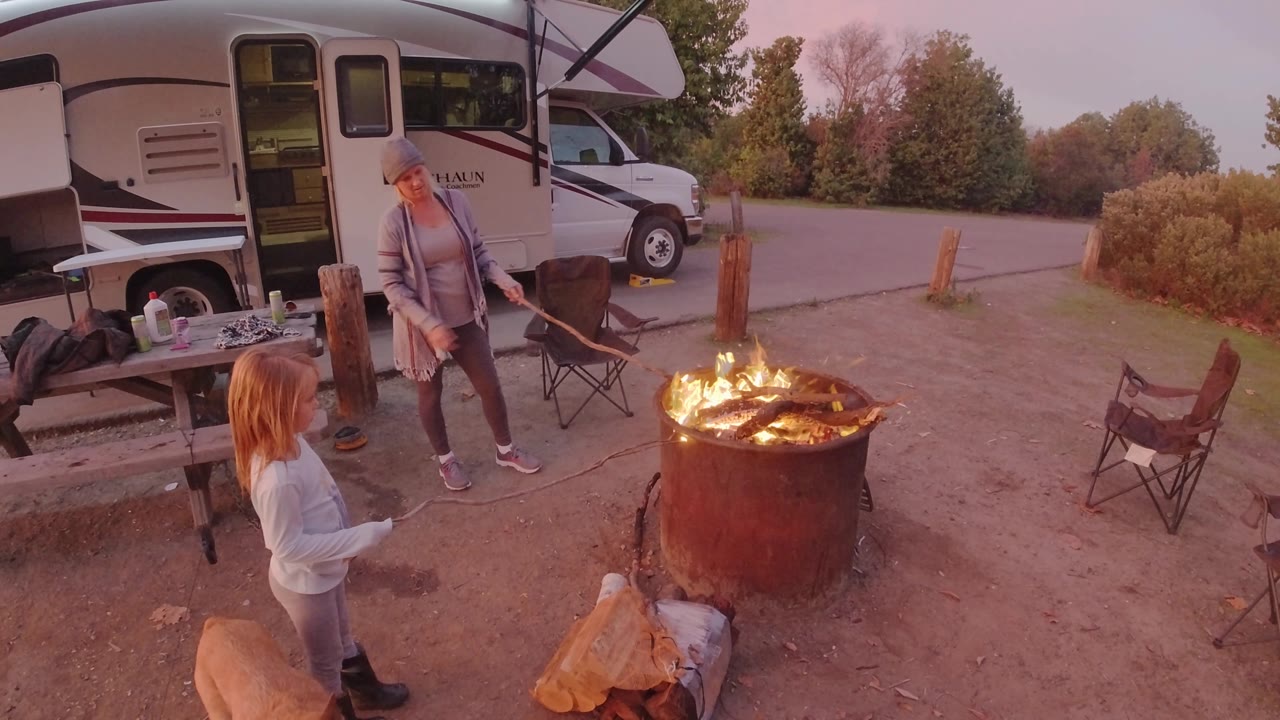 The child stands closer to the foreground while the adult leans slightly near the fire ring, with the RV side, open camp area, empty chair, bushes, and tree line in view.