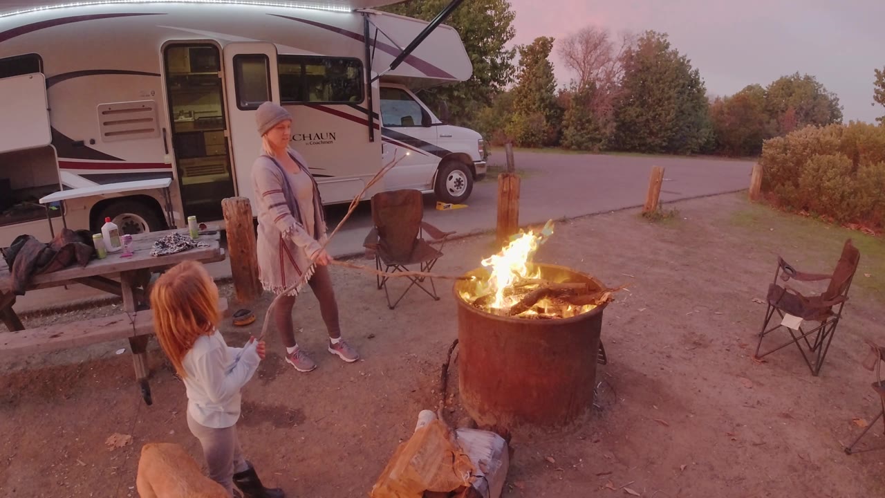 At 0:02, the young girl lifts a stick toward the woman as flames rise from the metal fire ring, with the RV and open campsite area behind them in San Clemente.