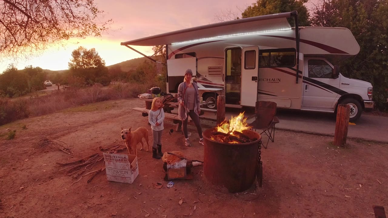In San Clemente, California, a woman, child, and dog stand near a flaming metal fire ring beside an RV under warm sunset light.