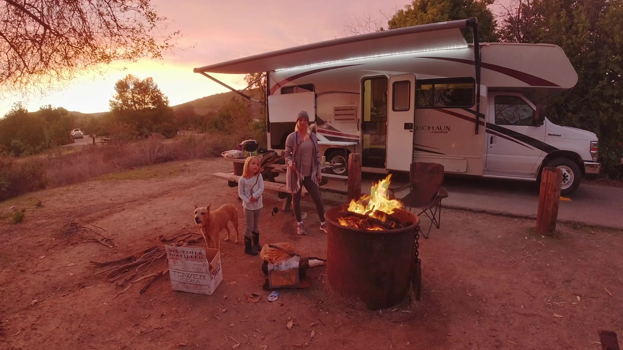 Child and dog stand near a fire ring while a woman is by the RV.