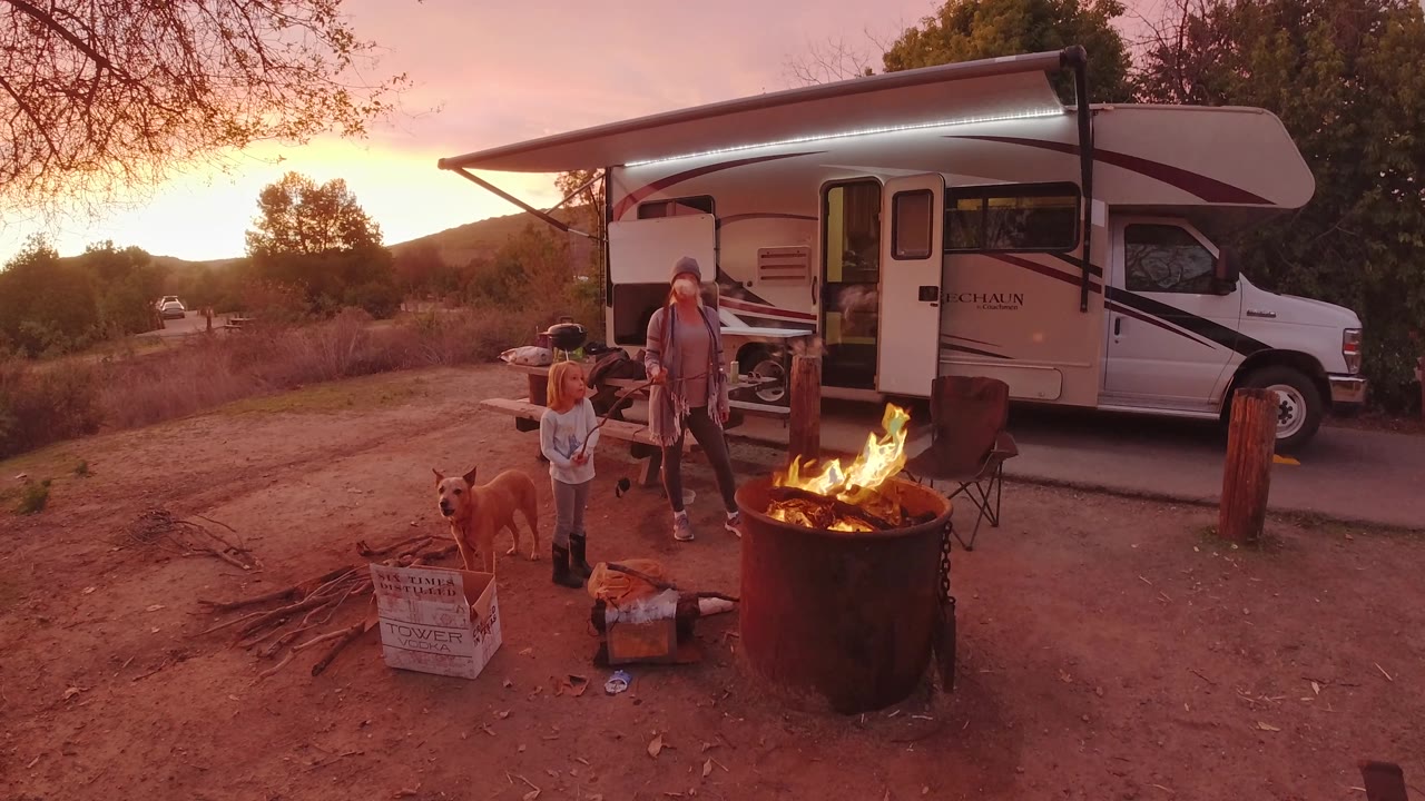 Flames rise higher in the foreground fire ring while a woman, child, and dog stand between the fire and the parked RV in San Clemente, California.