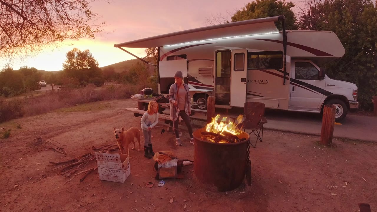 In San Clemente, California, a grey barrel fire burns in front of a white camper while two people and a dog stand near chairs and stacked wood.