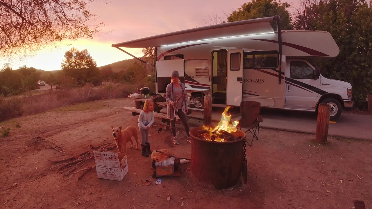 In San Clemente, California, the barrel fire flares in the foreground while two people, a dog, chairs, and the side of the white camper fill the campsite view.