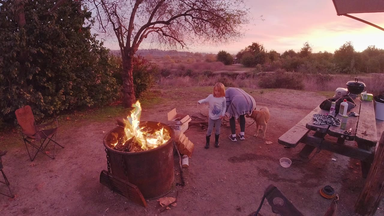 In San Clemente, California, a woman and child stand near a large metal fire pit as flames rise in the foreground and a picnic table sits to the right.