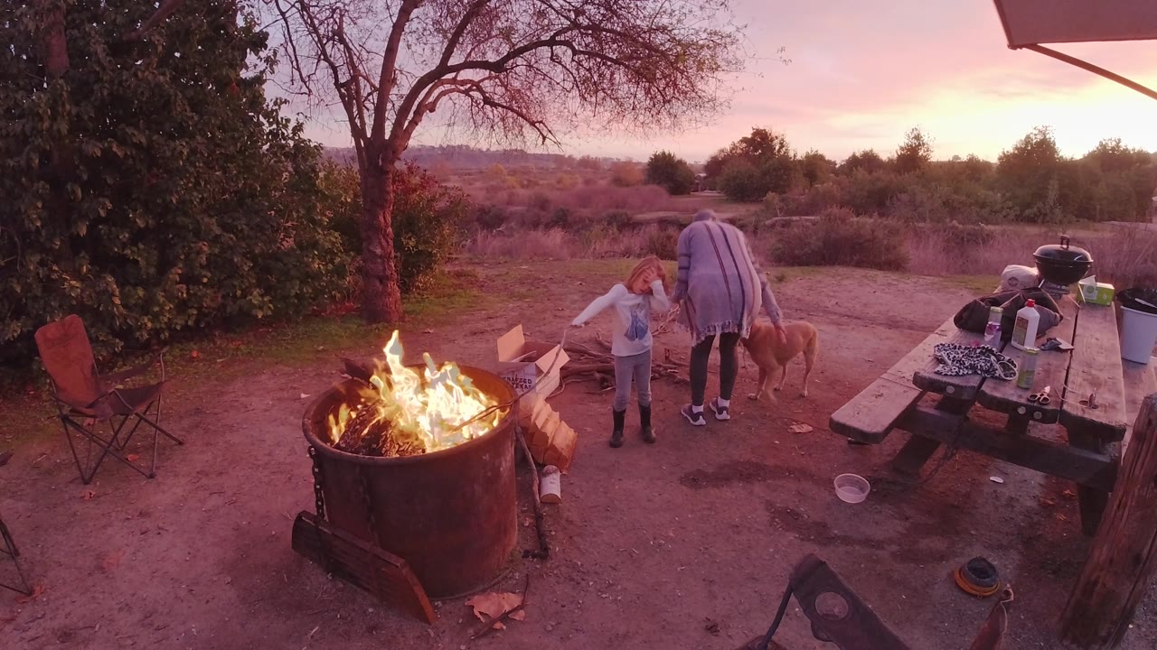 The adult stands over the child and tan dog near the right side of the fire pit, with a grill and picnic table visible under the pink sunset sky in San Clemente.