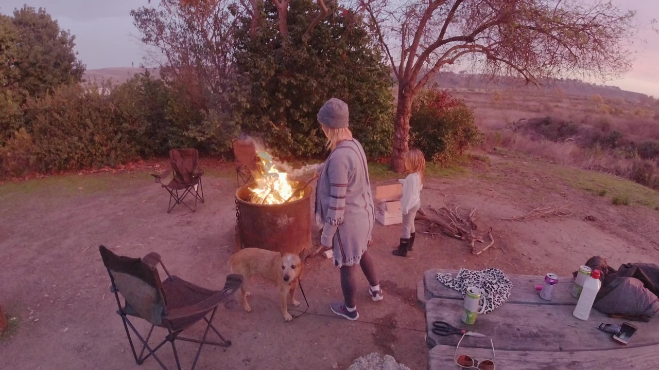 Elevated view in San Clemente, California of an adult in a knit cap, a blond child, and a tan dog standing near a bright metal fire barrel at the start of the clip.