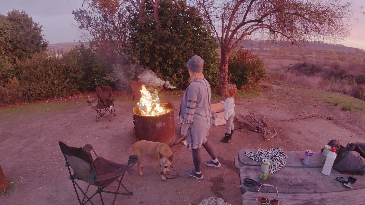 Adult, child, dog, chairs, and camp gear beside an open fire.