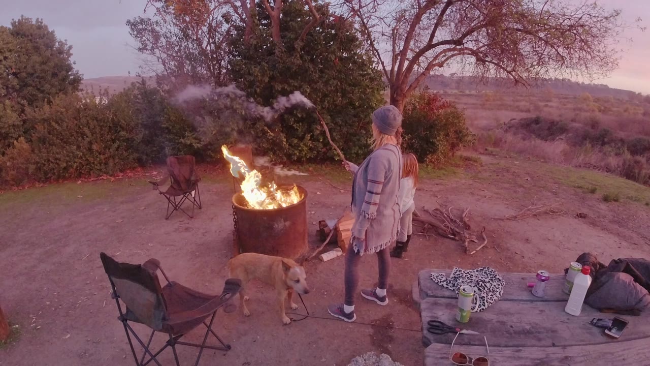 A woman and child stand beside a burning barrel campfire in San Clemente, California, with a tan dog, folding chairs, bags, and dusk hills in the background.