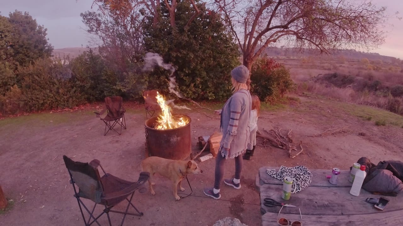 A bright flame rises from the metal barrel while the woman and child stand nearby in San Clemente, California, with chairs, loose firewood, and a tan dog in the campsite.