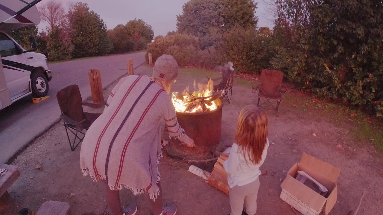 At 0:00 in San Clemente, a woman in a striped poncho leans toward a burning metal fire pit while a child stands beside logs and a cardboard box.