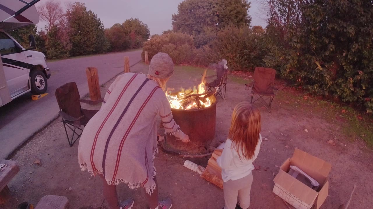 At 0:01 in San Clemente, the woman bends closer to the flames as the child faces the fire, with chairs, shrubs, logs, and the white van around the campsite-like setup.