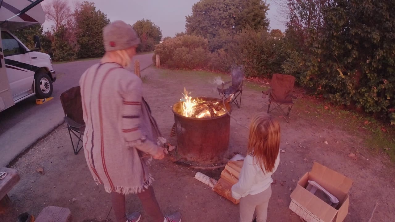 At 0:02 in San Clemente, the woman stands more upright with her back to camera while the child remains near the fire pit and smoke drifts over the chairs and shrubs.
