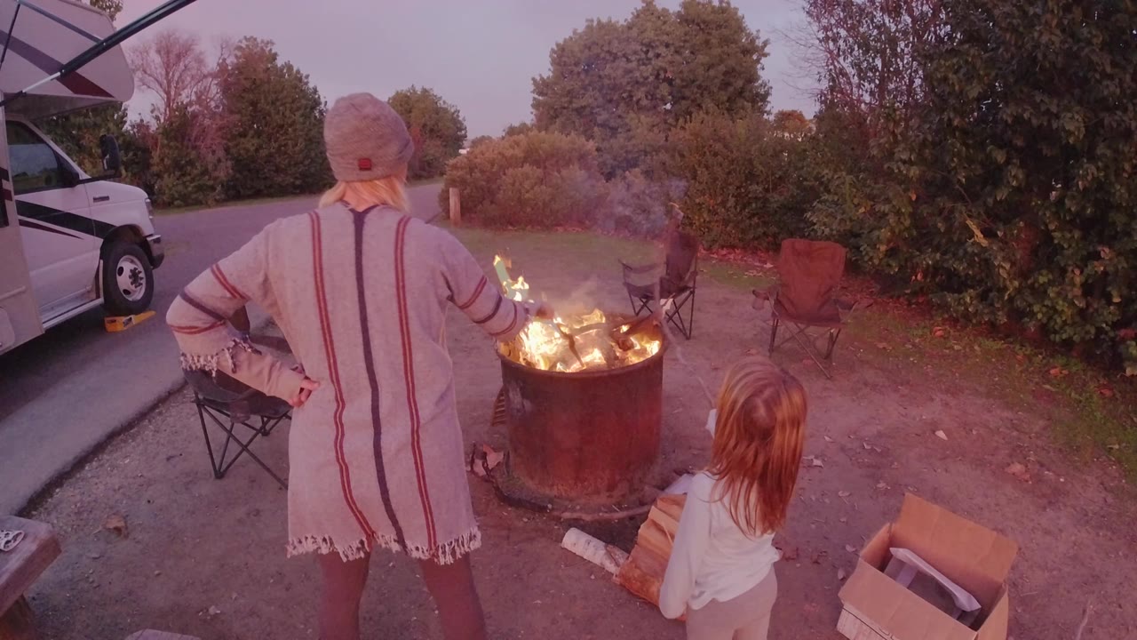 In San Clemente, California, a woman in a knit cap stands with her back to the camera while a child sits beside a barrel fire near folding chairs and a white vehicle.