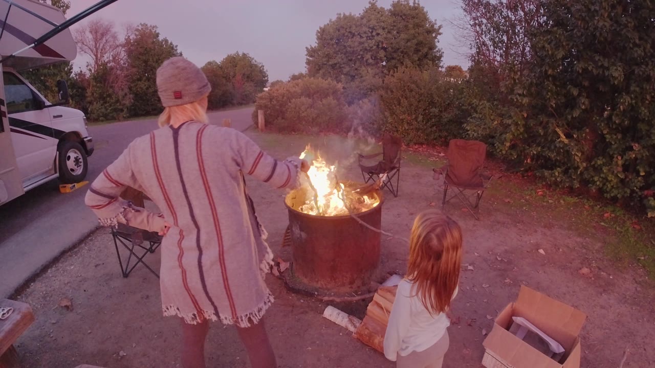In San Clemente, California, the woman extends one arm toward the burning barrel while the child sits close by, with folding chairs, shrubs, and a parked white vehicle around the roadside fire area.