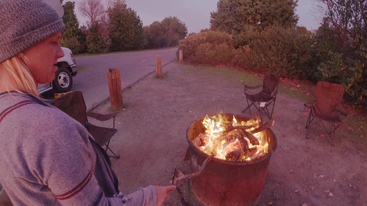 In San Clemente, California, a woman in a gray knit beanie stands at the left edge of the frame beside a round metal fire pit with bright flames and chairs behind it.
