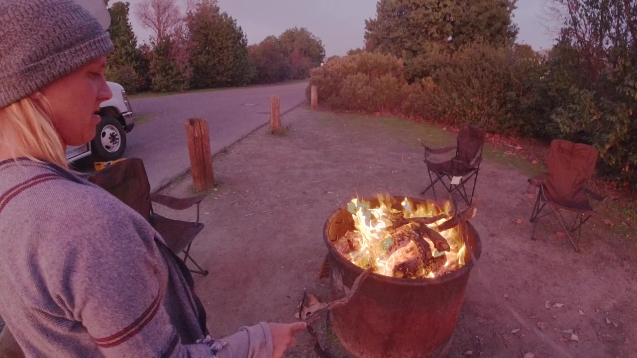 Woman holds a tool near a bright fire pit outdoors.
