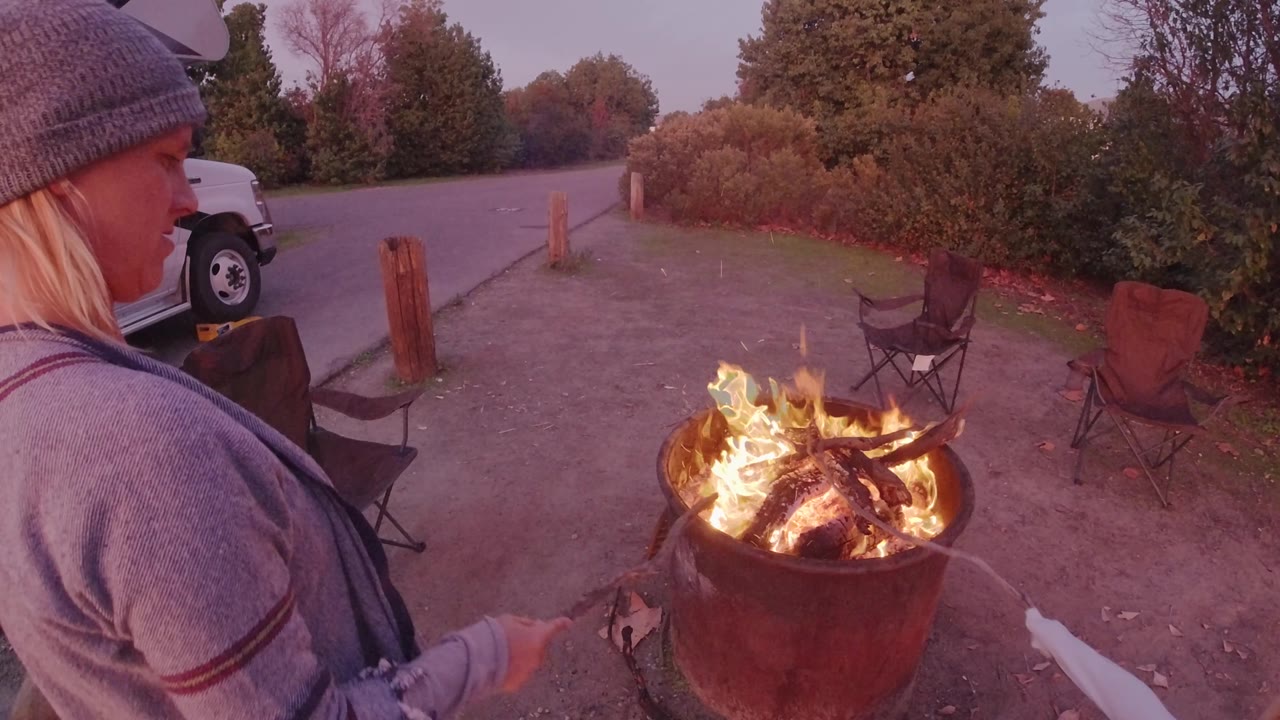 In San Clemente, California, the woman reaches farther toward the fire pit as flames flare above the logs and folding chairs sit on the gravel behind her.