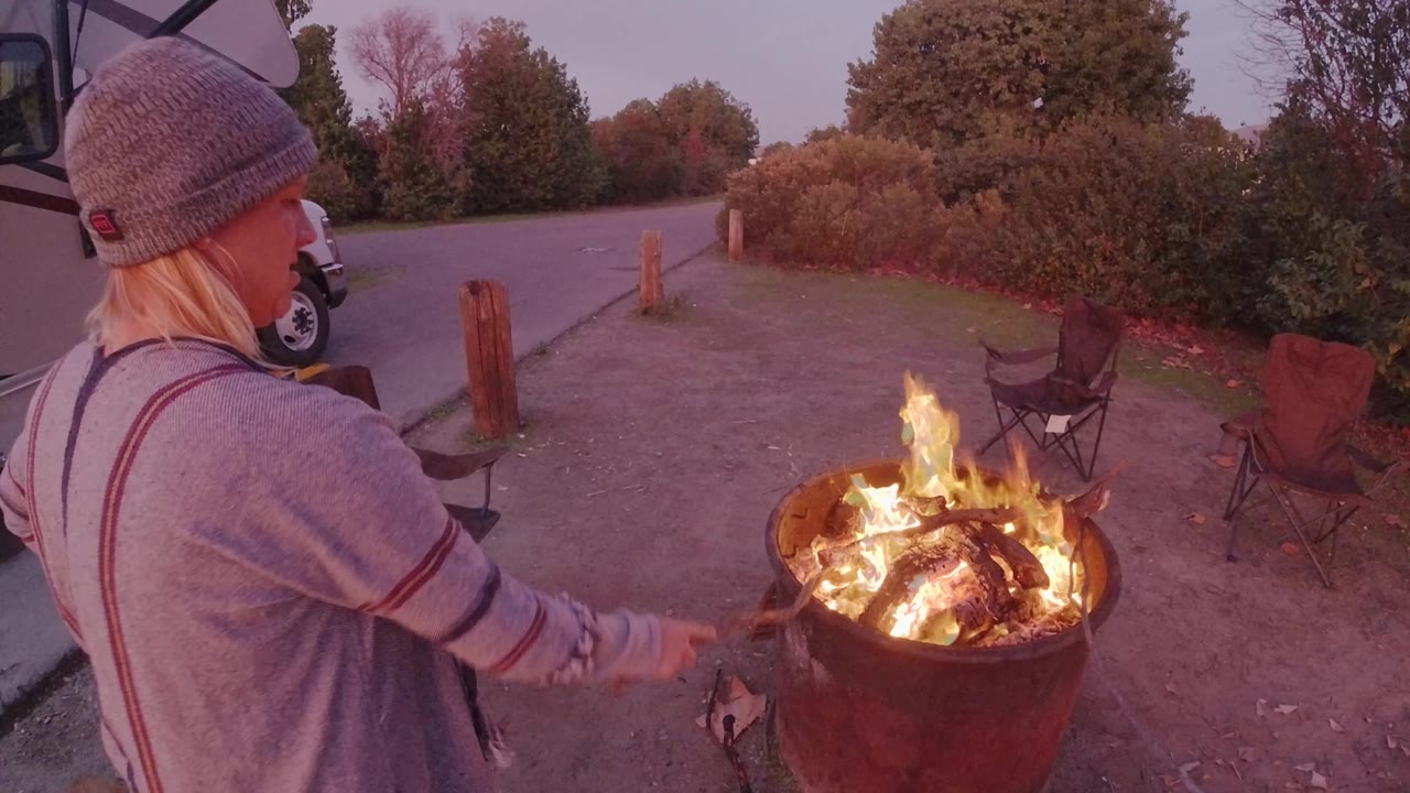 In San Clemente, California, an adult in a knit beanie stands at the left edge while reaching toward a metal fire pit burning beside folding camp chairs at dusk.