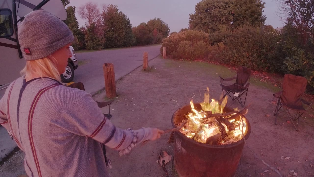 An adult camper van traveler in a gray knit hat stands beside a glowing fire pit in San Clemente, with empty folding chairs and roadside vegetation behind the flames.