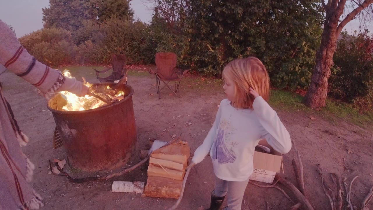 In San Clemente, California, a child stands beside a round metal fire pit as an adult arm reaches toward the flames, with split wood, sticks, chairs, and trees around the campsite.