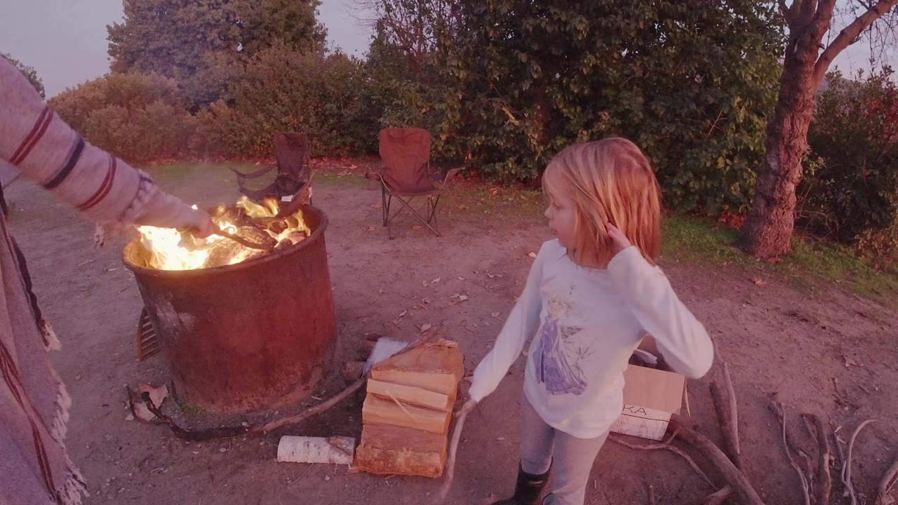 In San Clemente, California, an adult arm extends across the top of the frame toward the burning fire pit while the child stands on the right beside firewood and campsite gear.