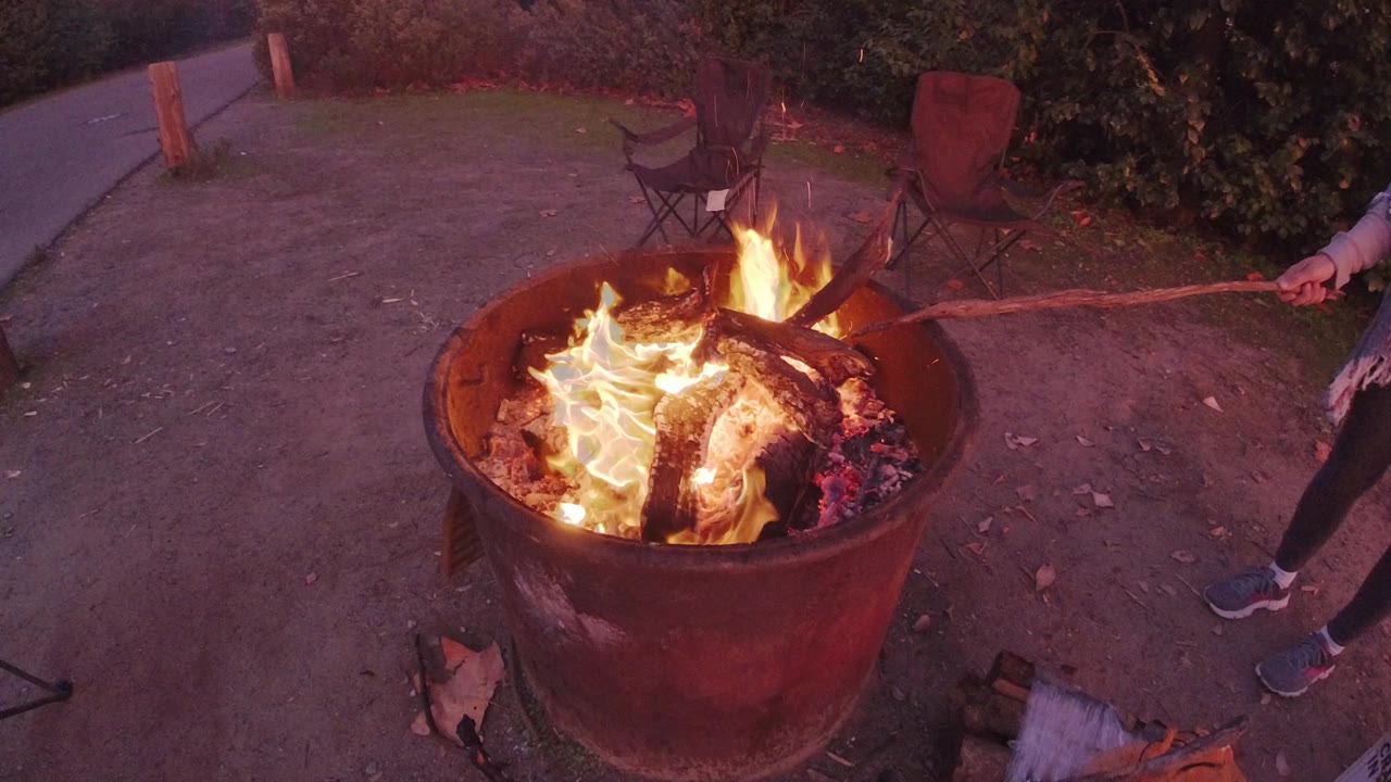 A metal barrel campfire burns in San Clemente, California, with bright flames around charred logs, folding chairs in the background, and a partially visible person at the right edge.