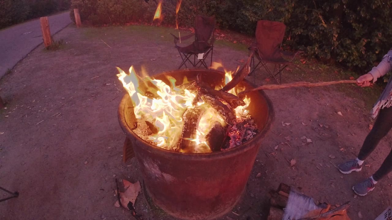 Flames flare higher across the left side of the metal barrel in San Clemente, California, while a partially visible person holds a long stick near the rim and chairs sit behind the fire.