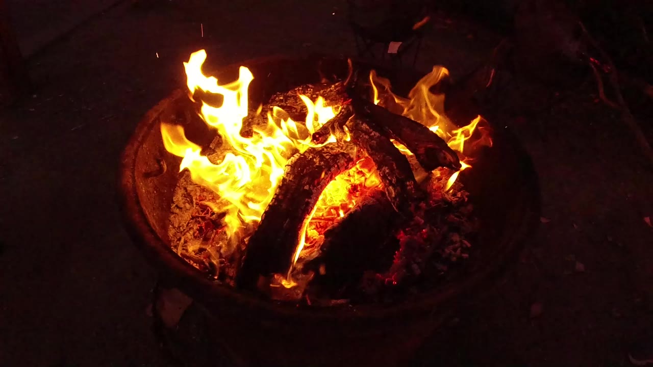 A high-angle view shows bright flames rising from charred logs in a round fire pit in San Clemente, California, with no visible people.