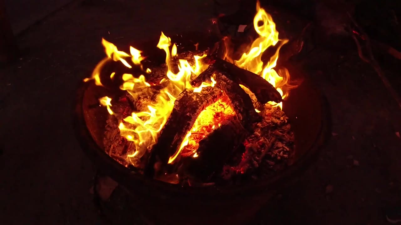 Tall yellow flames curl across blackened logs and ash in the fire pit, surrounded by darkness in San Clemente, California, with no visible people.