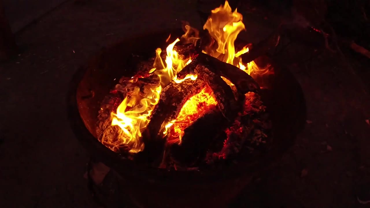 Flames concentrate along the left side of the charred logs while the ember bed glows in the San Clemente fire pit, with no visible people.