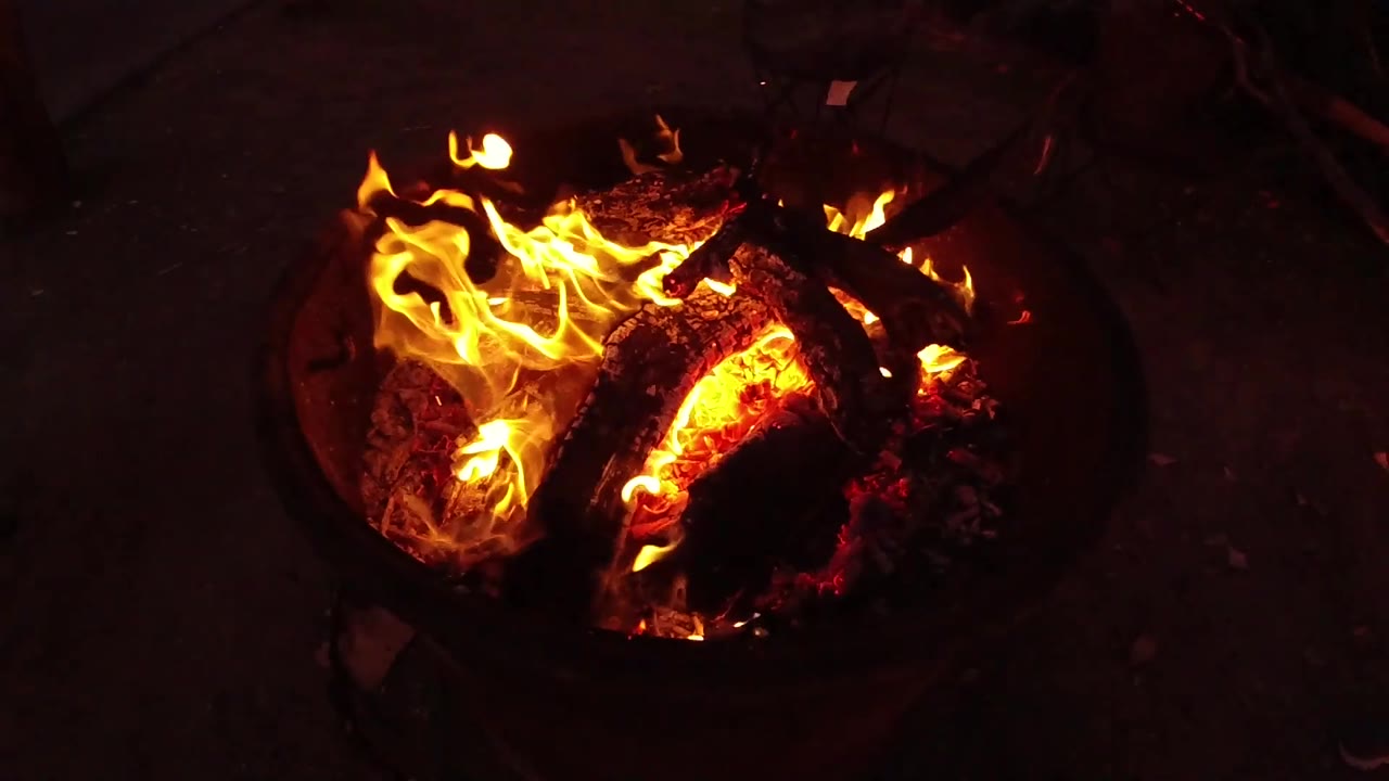 At 0:02, the fire spreads wider across the top of the logs, with bright orange flames and deep shadow around the pit, and no visible people.