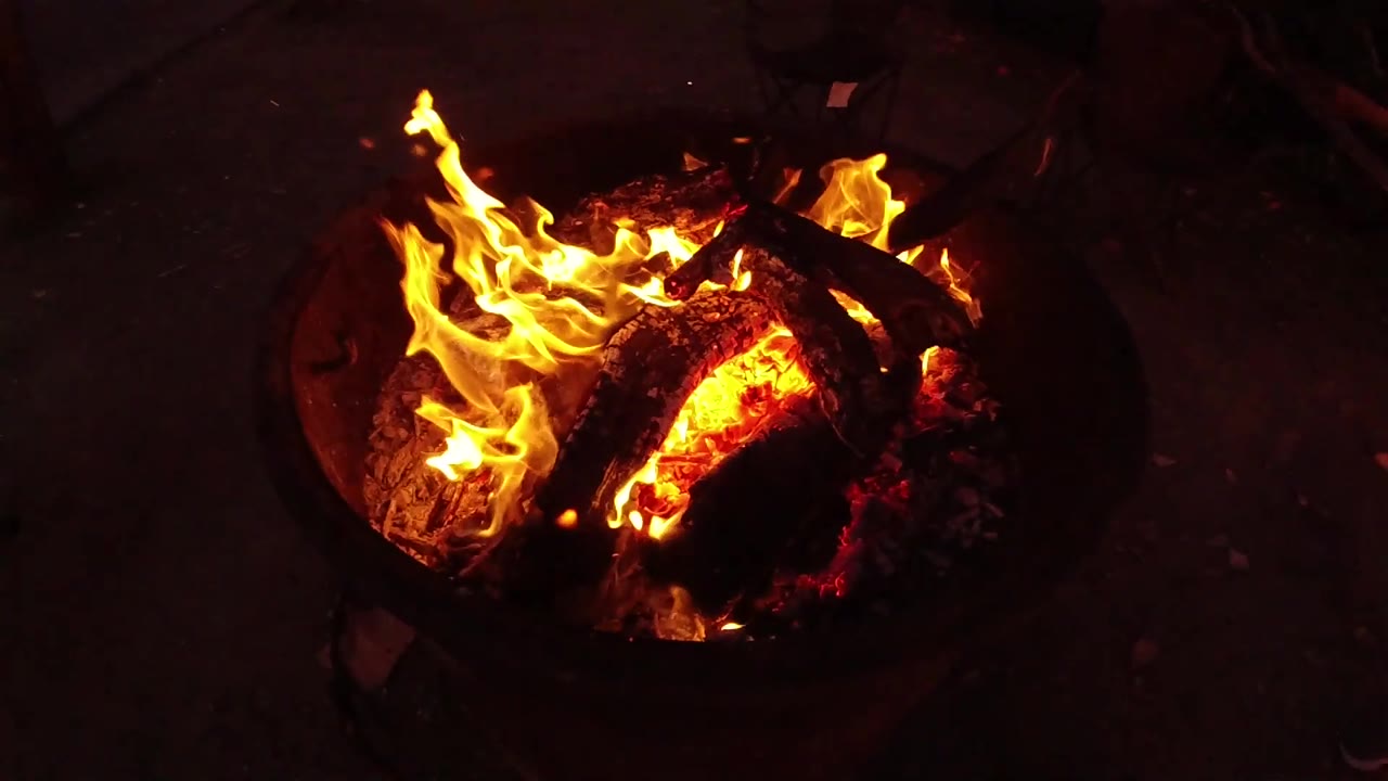 At 0:03, a taller flame rises on the left side of the round fire pit while red embers burn beneath the logs, with no visible people.
