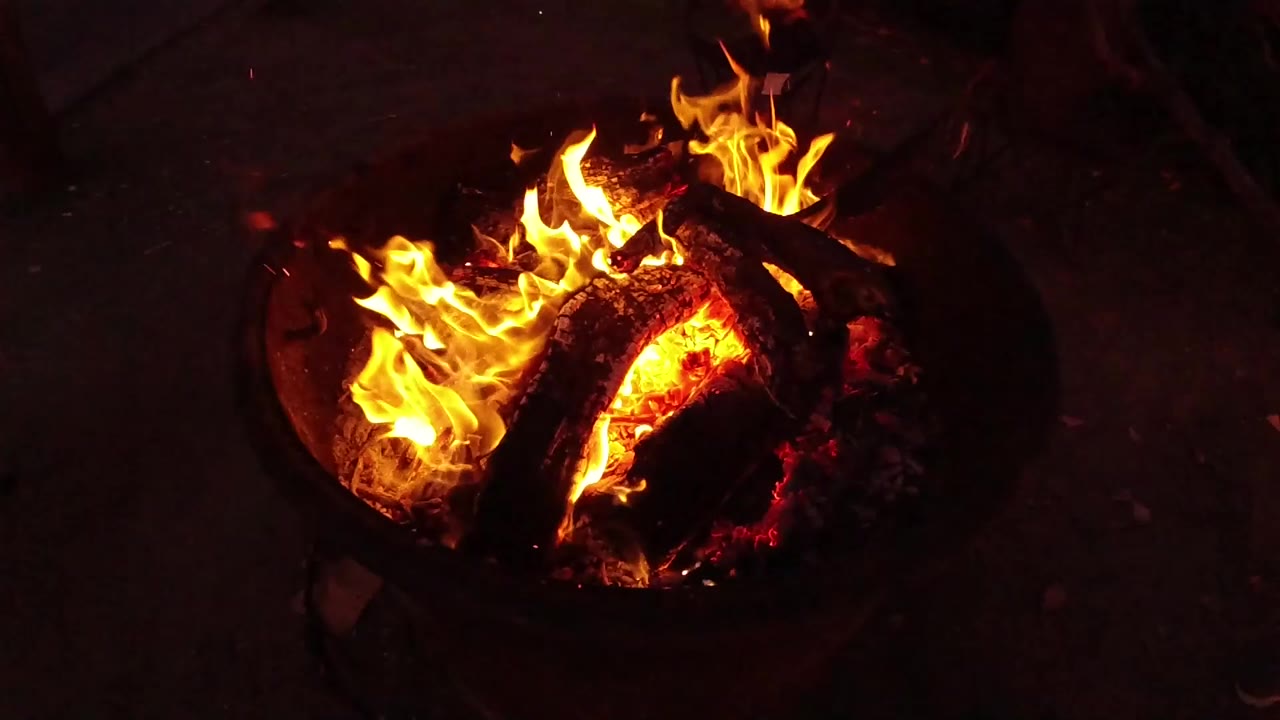 High-angle view of bright flames around charred logs in a dark fire pit from the San Clemente, California 2019 clip, with no visible people.