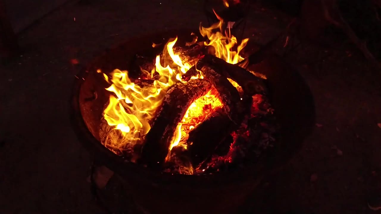 Flames climb higher over the left side of the charred wood in the round fire pit, with glowing embers visible and no visible people.
