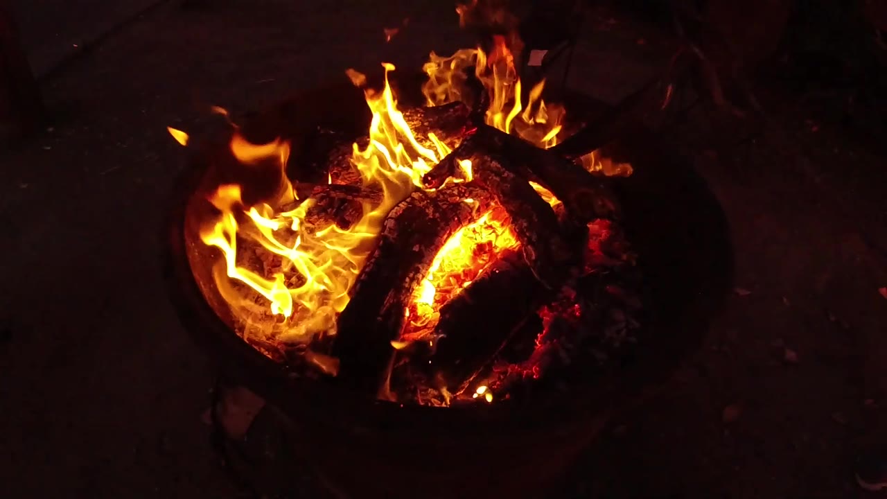 Yellow flames curl around blackened logs in the center of the fire pit, with red-orange heat visible through cracks and no visible people.