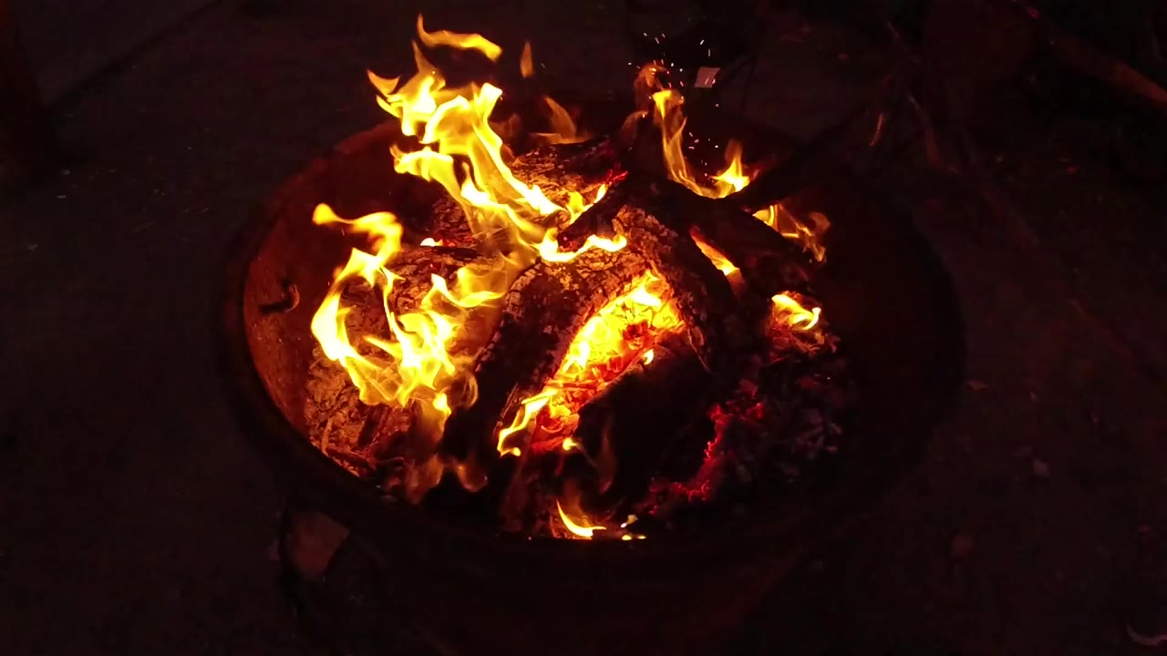 High-angle view of a round fire bowl in San Clemente, California, with orange flames wrapping around charred logs and no visible people.