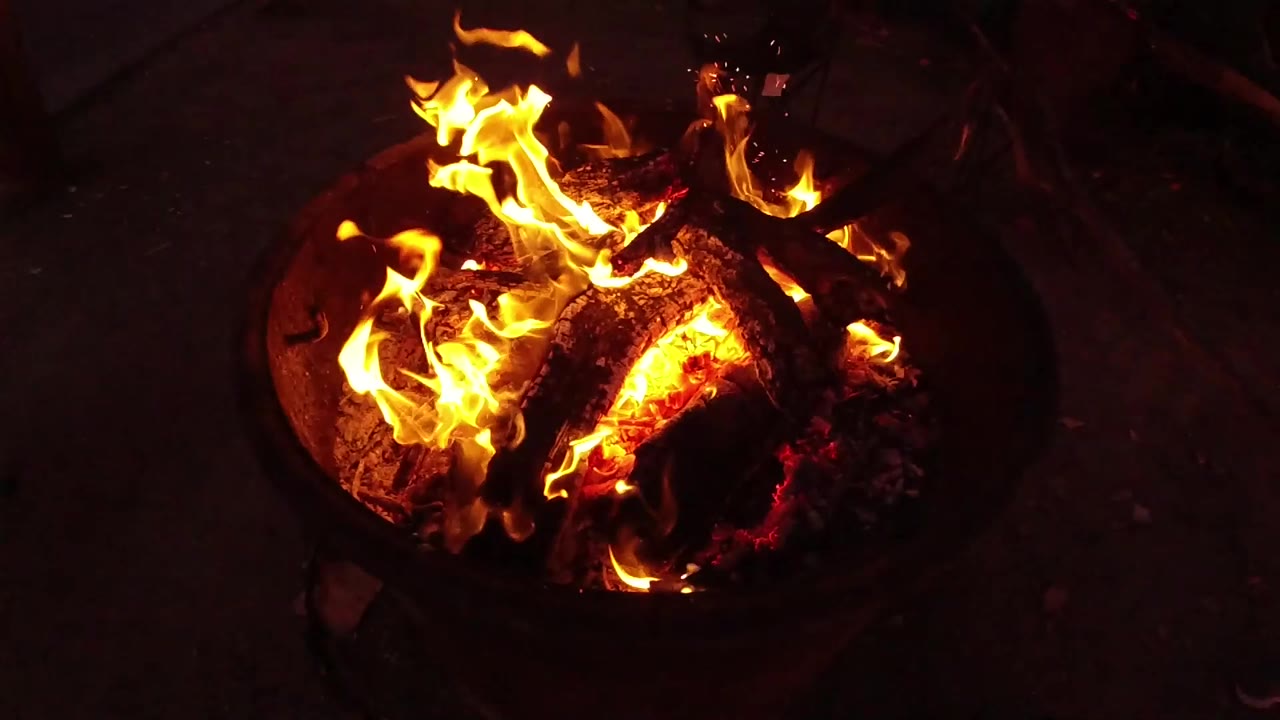 San Clemente night fire detail with flames leaning across blackened wood, red embers glowing below, and no visible people.