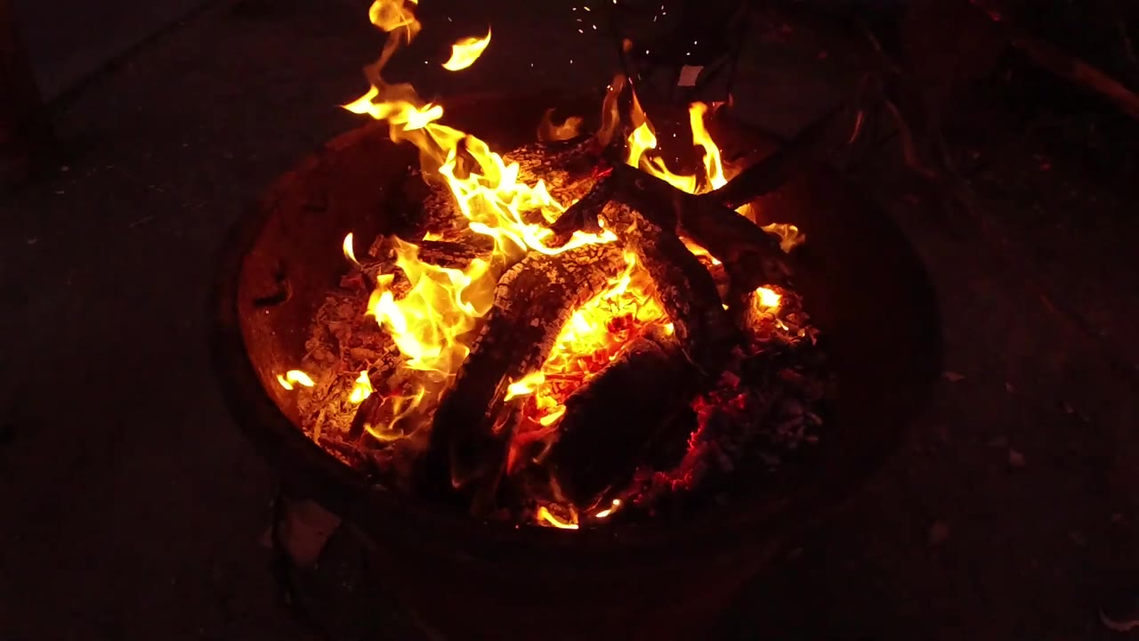 Round fire pit in the San Clemente collection with a taller flame flare on the upper left, glowing embers at the base, and no visible people.