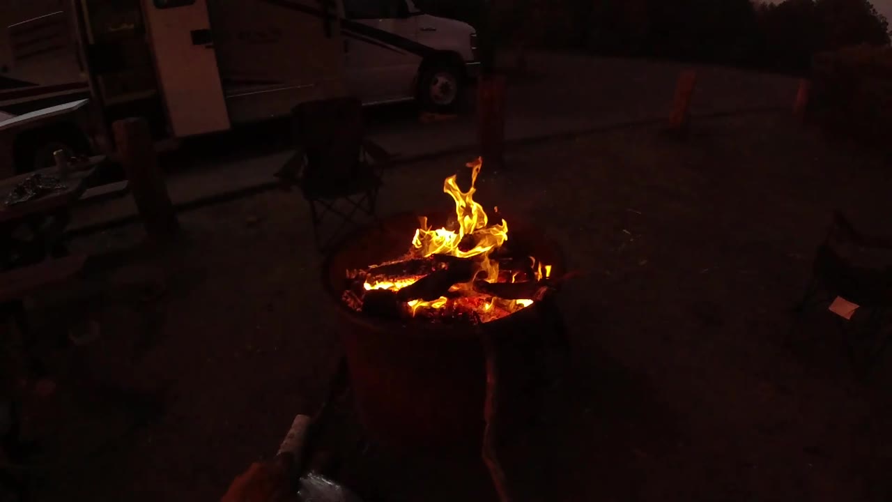 At 0:02, taller flames lift from the right side of the log stack in the metal fire pit, with dim pavement, a vehicle body, and no visible people in the background.