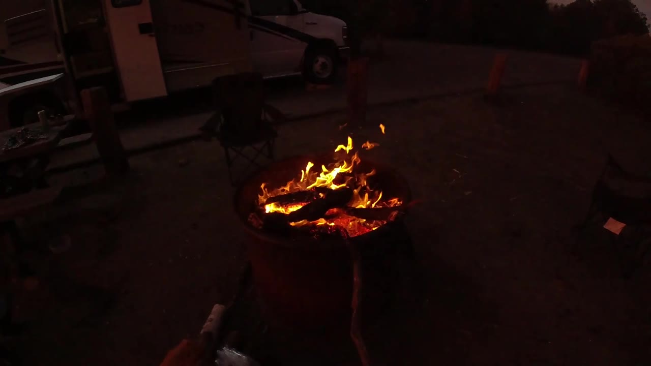 At 0:03, lower flames run along the log surfaces in the round fire pit, with red ember glow, dark open ground, background camping gear, and no visible people.
