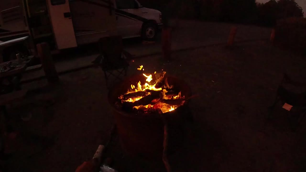 At 0:00 in San Clemente, California, a round fire pit glows with orange flames while a white van, chair, and dark pavement sit behind it.