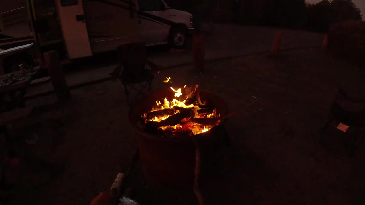 At 0:01 in San Clemente, California, the fire flares brighter over stacked logs, with the parked white van and chair still visible in the dark background.