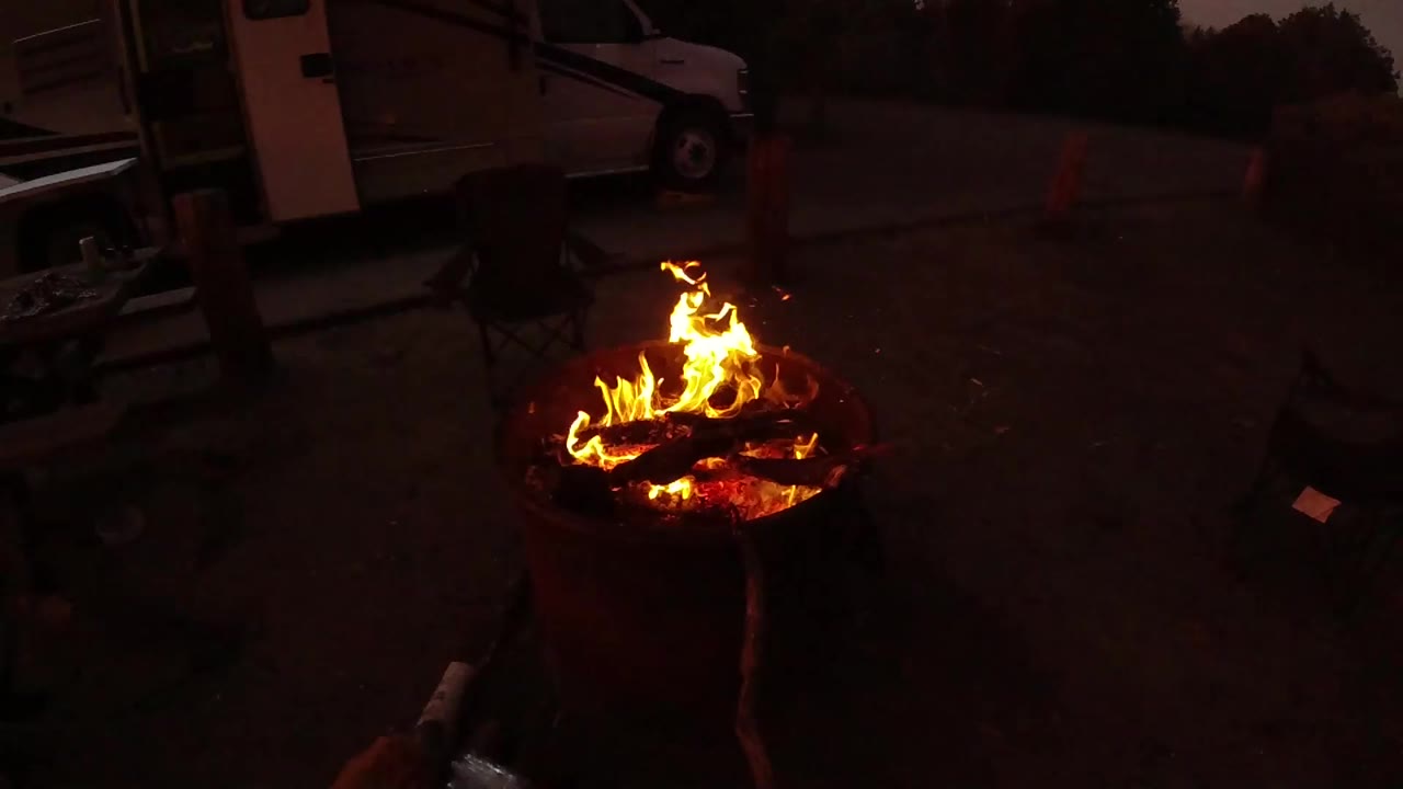 At 0:01, taller flames lift from the burning logs in the round fire pit, while the surrounding patio, chairs, and parked white vehicle or trailer stay dim and no people are visible.