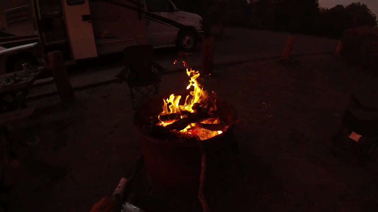 At 0:02, bright flames curl over glowing wood in the fire pit, with the dark patio and background vehicle or trailer visible behind it in San Clemente and no visible people.