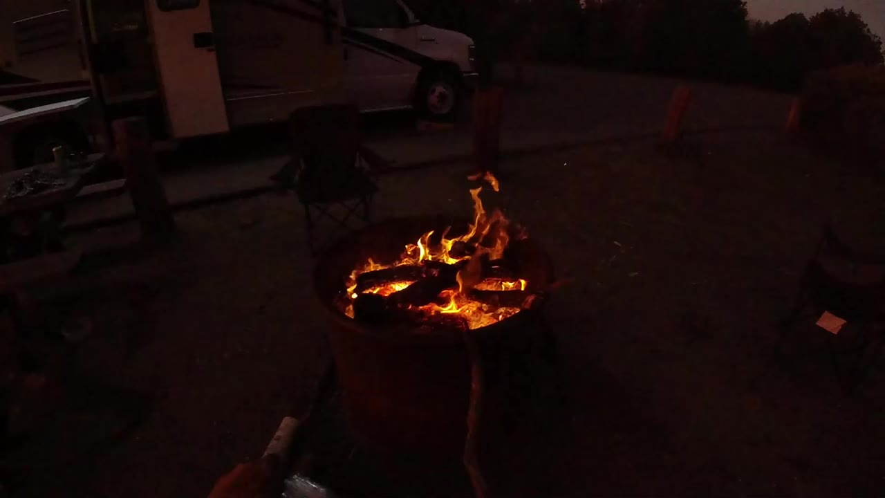 At 0:00, a round fire pit burns in a dark outdoor parking area in San Clemente, California, with a white truck or camper and folding chairs behind it.