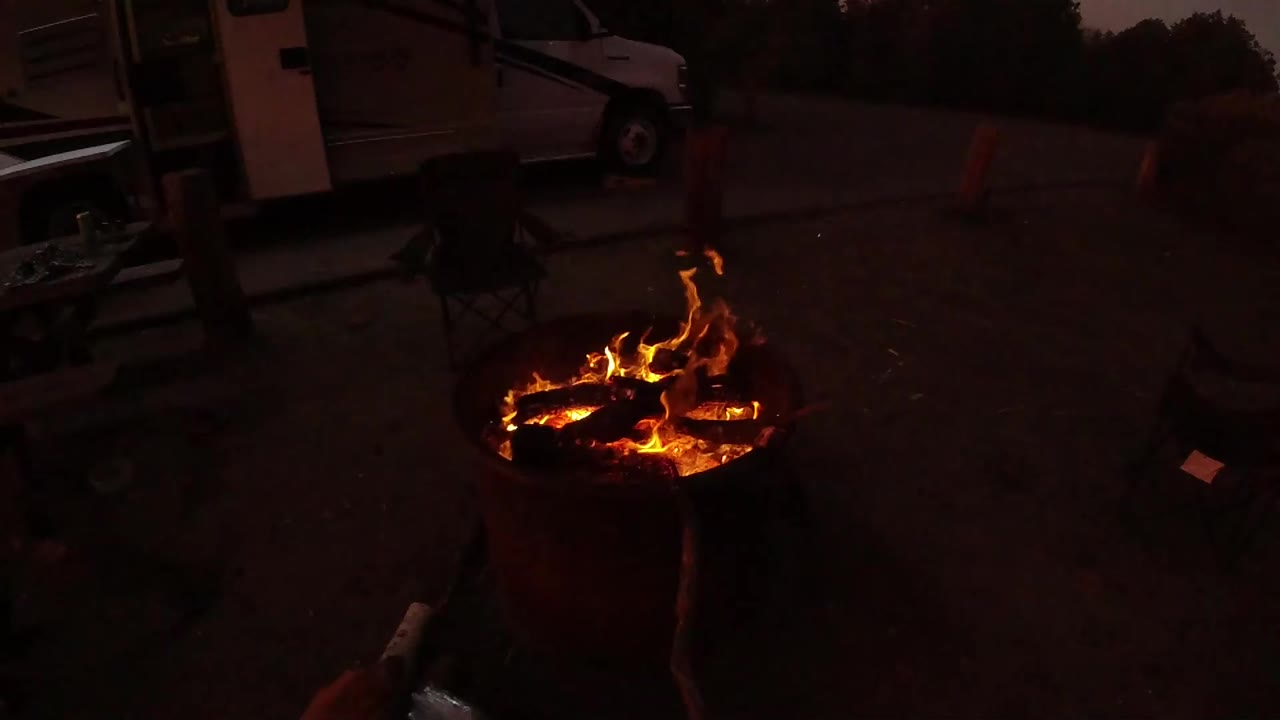 Bright flames rising from a fire pit with chairs and vehicles in shadow.