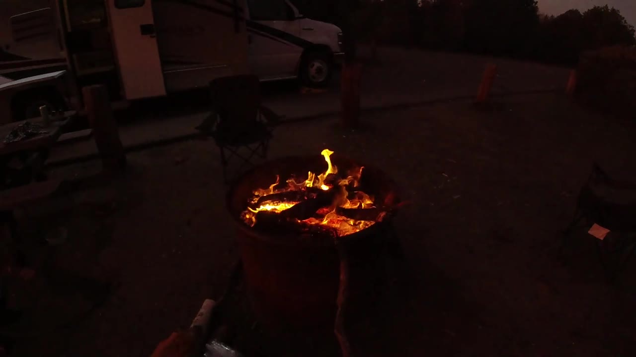 At 0:02, lower flames spread across the logs in the round pit, with an open dark lot, parked vehicle, and traffic cones visible behind it in San Clemente, California.