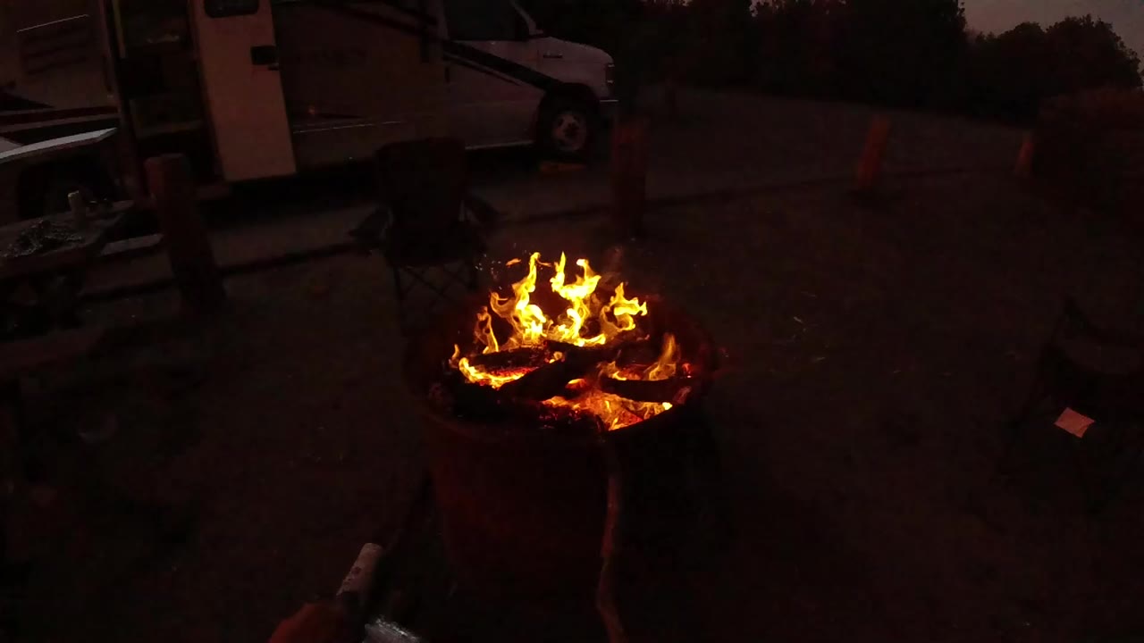 Tall flames rise from the burn barrel in the foreground while a dim parked vehicle, scattered campsite items, traffic cones, and a dark tree line sit behind it in San Clemente.