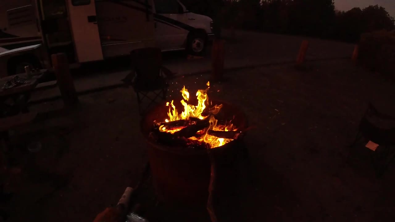 The burn barrel fire burns hot at the lower right of the frame, with a shadowed campsite driveway, parked vehicle, cones, trees, and an indistinct figure in the background.
